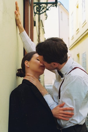 A couple is intimately embracing and kissing in a narrow alley with light-colored walls and an ornate street lamp overhead. The man is wearing a white shirt with suspenders, while the woman is dressed in a black jacket. The setting appears to be an urban environment, possibly in a historic part of a city.