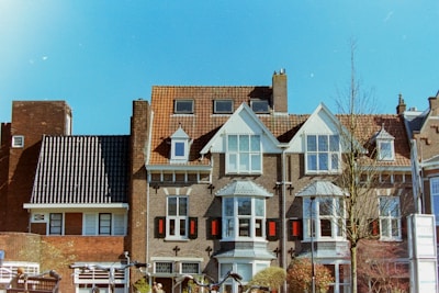 A row of European-style brick houses with white-framed windows, steeply pitched roofs, and ornate architectural details. The bright blue sky adds contrast to the warm tones of the buildings. Trees and greenery surround the area, with some bare branches visible, suggesting a cool season.
