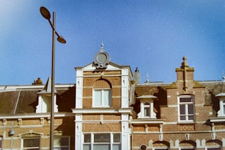 An architectural scene featuring a vintage European-style building with intricate brickwork and decorative elements. The building includes several windows with white framing, and the roof is adorned with ornate stonework. A tall streetlamp stands prominently in the foreground against a clear blue sky.