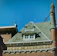 A rooftop of a historic building with ornate architectural details, featuring a steeply pitched roof with green shingles. The facade includes decorative brickwork and stone elements, with a distinctive chimney adorned with sculptural features. A small window near the roof adds character to the composition, while the deep blue sky provides a contrasting backdrop.