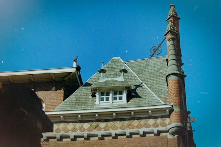 A rooftop of a historic building with ornate architectural details, featuring a steeply pitched roof with green shingles. The facade includes decorative brickwork and stone elements, with a distinctive chimney adorned with sculptural features. A small window near the roof adds character to the composition, while the deep blue sky provides a contrasting backdrop.