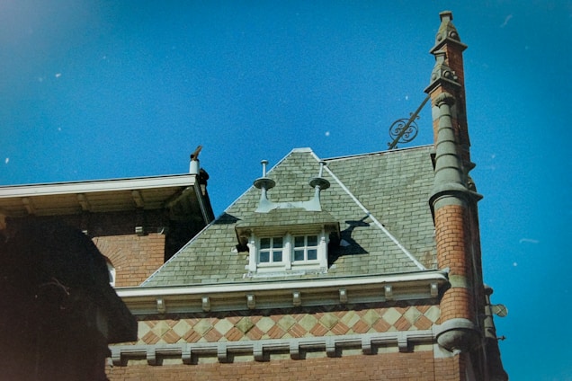 A rooftop of a historic building with ornate architectural details, featuring a steeply pitched roof with green shingles. The facade includes decorative brickwork and stone elements, with a distinctive chimney adorned with sculptural features. A small window near the roof adds character to the composition, while the deep blue sky provides a contrasting backdrop.