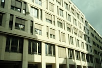 The image features a modern office building with a facade dominated by windows and rectangular patterns. The building's light exterior gives off reflections and shadows, indicating a sunny day. Several floors are visible, and the windows have blinds that are partially closed.