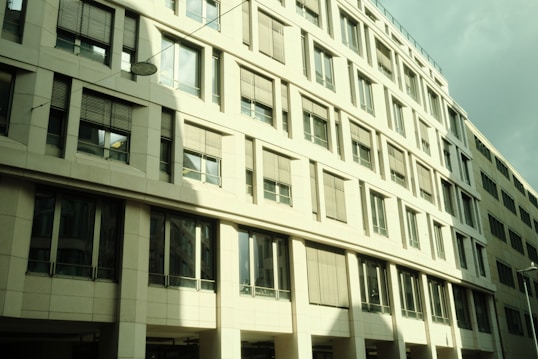 The image features a modern office building with a facade dominated by windows and rectangular patterns. The building's light exterior gives off reflections and shadows, indicating a sunny day. Several floors are visible, and the windows have blinds that are partially closed.