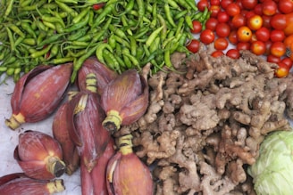 An assortment of fresh vegetables placed together, including green chilies, tomatoes, ginger, banana flower blossoms, and cabbage. The array showcases a variety of textures and vibrant colors.