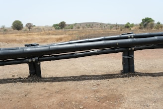 A dry landscape with brown grass and a few scattered trees is in the background. In the foreground, there are large black pipes elevated on supports, stretching horizontally across the image.