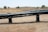 A dry landscape with brown grass and a few scattered trees is in the background. In the foreground, there are large black pipes elevated on supports, stretching horizontally across the image.