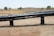 A dry landscape with brown grass and a few scattered trees is in the background. In the foreground, there are large black pipes elevated on supports, stretching horizontally across the image.