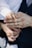 A close-up of a small child's hand gently placed on top of an adult's hand, which is wearing a gold band. The adult is dressed in a dark suit with a white shirt, and the child's sleeve is also white. The background is softly blurred, drawing focus to the hands.
