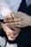 A close-up of a small child's hand gently placed on top of an adult's hand, which is wearing a gold band. The adult is dressed in a dark suit with a white shirt, and the child's sleeve is also white. The background is softly blurred, drawing focus to the hands.