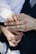 A close-up of a small child's hand gently placed on top of an adult's hand, which is wearing a gold band. The adult is dressed in a dark suit with a white shirt, and the child's sleeve is also white. The background is softly blurred, drawing focus to the hands.
