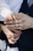 A close-up of a small child's hand gently placed on top of an adult's hand, which is wearing a gold band. The adult is dressed in a dark suit with a white shirt, and the child's sleeve is also white. The background is softly blurred, drawing focus to the hands.