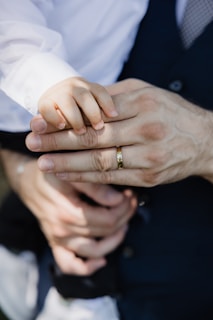A close-up of a small child's hand gently placed on top of an adult's hand, which is wearing a gold band. The adult is dressed in a dark suit with a white shirt, and the child's sleeve is also white. The background is softly blurred, drawing focus to the hands.