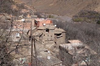 A rustic village is nestled within a mountainous landscape, featuring traditional mud-brick buildings, many of which have white satellite dishes on their rooftops. Bare tree branches are visible, indicating a season like winter or early spring. The backdrop comprises rolling hills or mountains, with a sparse covering of vegetation.