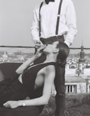 A woman reclining on a chair with elegant attire is held by a man standing next to her, lighting her cigarette. The scene is set against a cityscape backdrop. The woman wears a sleeveless dress and a wristwatch, while the man sports a bow tie, white shirt, and suspenders.