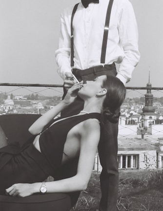 A woman reclining on a chair with elegant attire is held by a man standing next to her, lighting her cigarette. The scene is set against a cityscape backdrop. The woman wears a sleeveless dress and a wristwatch, while the man sports a bow tie, white shirt, and suspenders.