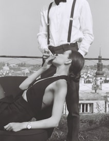 A woman reclining on a chair with elegant attire is held by a man standing next to her, lighting her cigarette. The scene is set against a cityscape backdrop. The woman wears a sleeveless dress and a wristwatch, while the man sports a bow tie, white shirt, and suspenders.