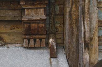 A close-up view of an aged, rustic wooden structure with visible weathering. The surface features intricate carvings and patterns, complemented by a rusted metal pipe extending downwards. The wood displays signs of peeling paint and natural decay, lending an antique and historic feel.
