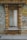An ornate wooden window frame with intricate carvings and a weathered, rustic appearance. The window is divided into two sections and features white geometric metal bars. The surrounding wall has peeling paint, adding to the antique charm.