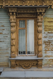 An ornate wooden window frame with intricate carvings and a weathered, rustic appearance. The window is divided into two sections and features white geometric metal bars. The surrounding wall has peeling paint, adding to the antique charm.