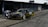 A sleek black race car with yellow accents and the number 25 is stationed in front of a garage. Two individuals are inspecting or preparing the car, with one person kneeling near the driver's side. The setting includes multiple garage doors, some of which are partially open, showing equipment and other racing gear.