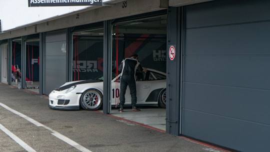 A person in a racing suit stands next to a white sports car with the number 10 on the side, in what appears to be a garage or pit lane. The garage door is open, and the indoor space contains various racing-related equipment. The exterior area shows concrete pavement with markings.