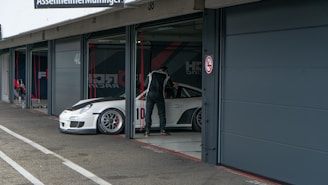 A person in a racing suit stands next to a white sports car with the number 10 on the side, in what appears to be a garage or pit lane. The garage door is open, and the indoor space contains various racing-related equipment. The exterior area shows concrete pavement with markings.