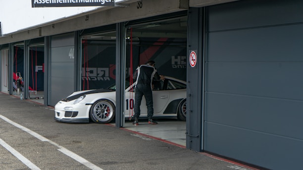 A person in a racing suit stands next to a white sports car with the number 10 on the side, in what appears to be a garage or pit lane. The garage door is open, and the indoor space contains various racing-related equipment. The exterior area shows concrete pavement with markings.