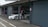 A person in a racing suit stands next to a white sports car with the number 10 on the side, in what appears to be a garage or pit lane. The garage door is open, and the indoor space contains various racing-related equipment. The exterior area shows concrete pavement with markings.