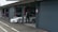 A person in a racing suit stands next to a white sports car with the number 10 on the side, in what appears to be a garage or pit lane. The garage door is open, and the indoor space contains various racing-related equipment. The exterior area shows concrete pavement with markings.