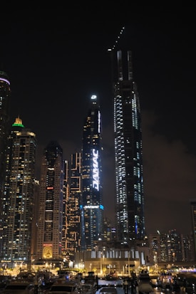A nighttime cityscape featuring tall, illuminated skyscrapers with bright signage in an urban environment. The skyline is densely packed and there's a marina in the foreground with several boats docked.