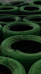A collection of stacked green tires arranged in a pattern, each with visible worn treads and weathered texture. The arrangement gives a repetitive yet organized appearance, suitable for an outdoor setting or a racing track barrier.