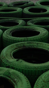 A collection of stacked green tires arranged in a pattern, each with visible worn treads and weathered texture. The arrangement gives a repetitive yet organized appearance, suitable for an outdoor setting or a racing track barrier.