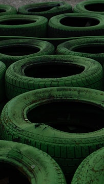 A collection of stacked green tires arranged in a pattern, each with visible worn treads and weathered texture. The arrangement gives a repetitive yet organized appearance, suitable for an outdoor setting or a racing track barrier.