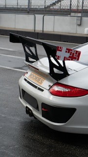 A white sports car with a prominent rear spoiler is parked on what appears to be a wet race track. The car has a 'ROOKIES' license plate and red accents on the rear lights. The spoiler is large and black with a red and white pattern on its sides.
