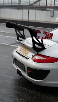 A white sports car with a prominent rear spoiler is parked on what appears to be a wet race track. The car has a 'ROOKIES' license plate and red accents on the rear lights. The spoiler is large and black with a red and white pattern on its sides.