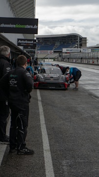 Several people in pit crew uniforms are gathered around a racing car in a pit lane, likely preparing it for a race. The setting appears to be a motorsport event, with advertisements for automotive services visible in the background. The weather is overcast, and the surface of the pit lane is wet, suggesting recent rain.