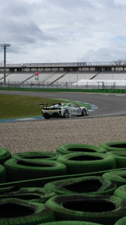 A racing car speeds through a circuit against a backdrop of empty stands and cloudy skies. The vehicle is decorated with advertising graphics and moves along a curved section of the track, which is lined with green tire barriers in the foreground.
