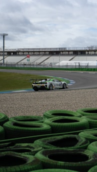 A racing car speeds through a circuit against a backdrop of empty stands and cloudy skies. The vehicle is decorated with advertising graphics and moves along a curved section of the track, which is lined with green tire barriers in the foreground.