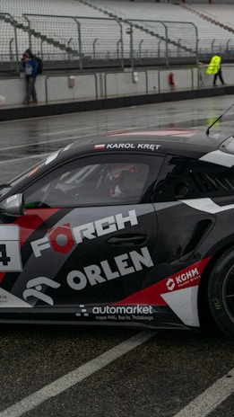 A black race car with the labels 'FORCH' and 'ORLEN' on its sides is parked on a wet racetrack. The car appears to be designed for motorsport, with a visible driver's name at the top of the window. Spectators and officials are present in the background, and the seating areas are mainly empty. The scene takes place in an overcast or rainy setting, indicated by the wet surface.