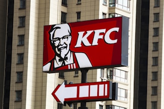 A KFC sign is prominently displayed with a red background, featuring the company's logo and an illustration of the founder. The sign is mounted on a pole, with a pointer indicating direction. Tall buildings with multiple windows are seen in the background.