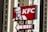 A KFC sign is prominently displayed with a red background, featuring the company's logo and an illustration of the founder. The sign is mounted on a pole, with a pointer indicating direction. Tall buildings with multiple windows are seen in the background.