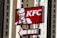 A KFC sign is prominently displayed with a red background, featuring the company's logo and an illustration of the founder. The sign is mounted on a pole, with a pointer indicating direction. Tall buildings with multiple windows are seen in the background.