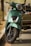 A close-up view of a green scooter parked on a street. The scooter features a modern design with a chrome front grille and a large headlight. It rests on its kickstand, and the scene is set in an outdoor environment with blurred trees and pavement in the background.