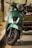 A close-up view of a green scooter parked on a street. The scooter features a modern design with a chrome front grille and a large headlight. It rests on its kickstand, and the scene is set in an outdoor environment with blurred trees and pavement in the background.