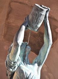 A bronze statue of a person holding a barrel above their head, with liquid pouring from the barrel into their mouth. The figure's arms are raised and the expression is neutral. The background features a stone arch with warm hues.