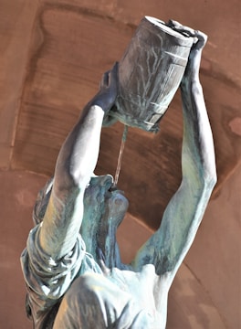 A bronze statue of a person holding a barrel above their head, with liquid pouring from the barrel into their mouth. The figure's arms are raised and the expression is neutral. The background features a stone arch with warm hues.