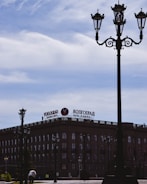 A historic building with an architectural design is visible, featuring large windows and a prominent sign on top that reads 'Volgograd' in Cyrillic and English. Several ornate street lamps are positioned in the foreground, with a cloudy sky above.