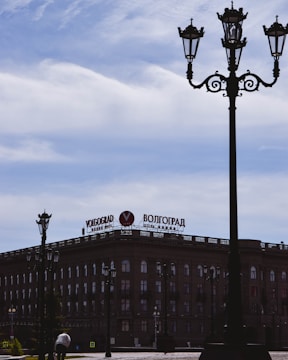 A historic building with an architectural design is visible, featuring large windows and a prominent sign on top that reads 'Volgograd' in Cyrillic and English. Several ornate street lamps are positioned in the foreground, with a cloudy sky above.