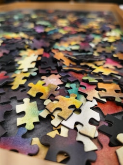 A collection of colorful jigsaw puzzle pieces scattered on a surface. The pieces display a variety of colors including shades of yellow, pink, green, and blue. The image focuses on the texture and complexity of the interlocking pieces, highlighting the randomness of their arrangement.